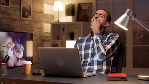 Manager stanco che sbadiglia mentre lavora al computer portatile in ufficio a casa durante le ore notturne. Uomo d'affari oberato di lavoro.