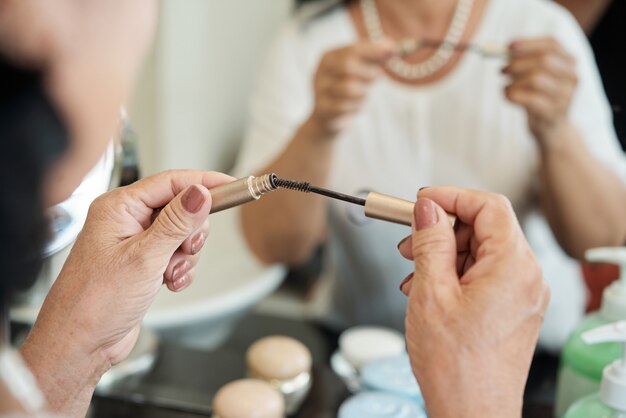 Mani del mascara senior irriconoscibile che tengono mascara davanti allo specchio