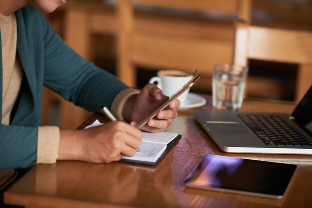 Mani dell'uomo irriconoscibile che si siede al tavolo nella caffetteria con gadget e la scrittura nel quaderno