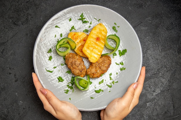 Mani della donna che tengono la cena gustosa con il pasto della cotoletta insieme alle patate sul piatto bianco sulla tavola nera