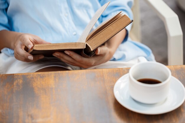 Mani della ragazza che si siedono al tavolo con la tazza di caffè e del libro.