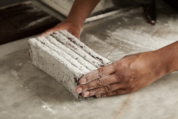 Mani dello chef uomo nero tiene barrette di cioccolato appena sfornate in polvere di zucchero prima del confezionamento, primo piano in pasticceria artigianale professionale