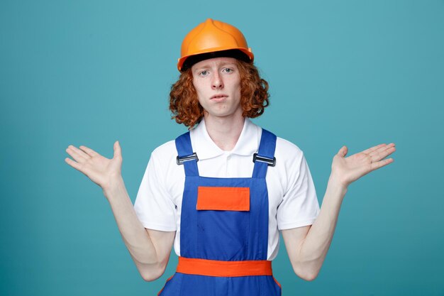 Mani di diffusione confuse giovane uomo costruttore in uniforme isolato su sfondo blu