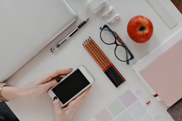 Mani di donna con smartphone alla scrivania con matite, occhiali e portatile