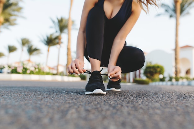 Mani di donna graziosa che allaccia i lacci delle scarpe su scarpe da ginnastica sulla strada con le palme. Mattina di sole in città tropicale, allenamento, fitness, allenamento, motivazione