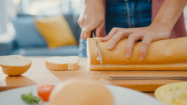 Mani di giovane donna asiatica chef azienda coltello taglio pane integrale sul bordo di legno