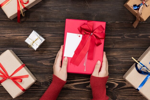 Mani femminili che tengono una piccola scatola con un regalo tra le decorazioni festive invernali su una vista da tavolo bianca. Composizione piatta per compleanno, natale o matrimonio.