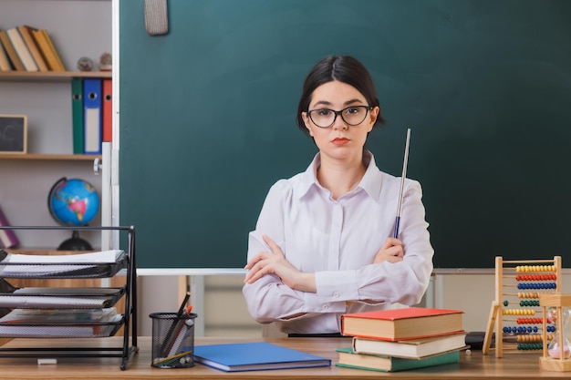 mani incrociate fiduciose giovane insegnante femminile che indossa occhiali tenendo il puntatore seduto alla scrivania con strumenti scolastici in classe