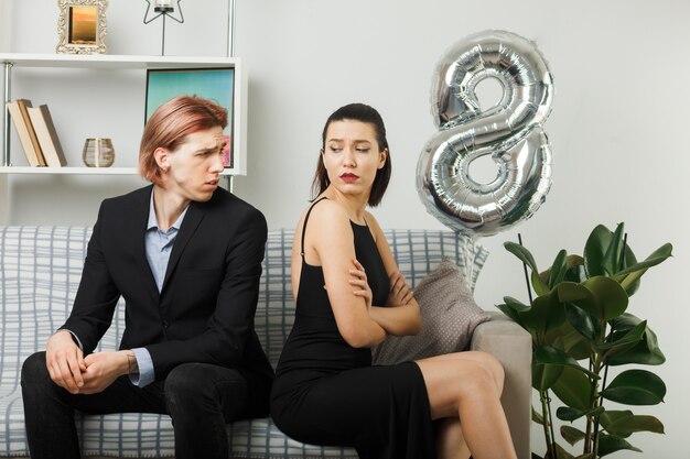Mani incrociate scontente di una giovane coppia in una felice giornata delle donne che si guardano seduti sul divano di schiena in soggiorno