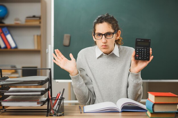 mani scontente che diffondono il giovane insegnante maschio che tiene la calcolatrice seduto alla scrivania con gli strumenti della scuola in classe