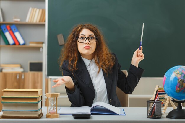 mani spalancate confuse giovane insegnante femminile che indossa occhiali punta alla lavagna con puntatore seduto alla scrivania con strumenti scolastici in classe