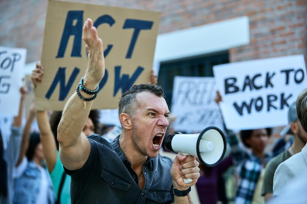 Manifestante arrabbiato che usa il megafono e grida mentre protesta con la folla di persone per le strade della città