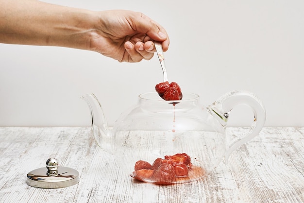 Mano che prende la marmellata di fragole con un cucchiaio dalla teiera di vetro su un tavolo di legno contro un muro bianco