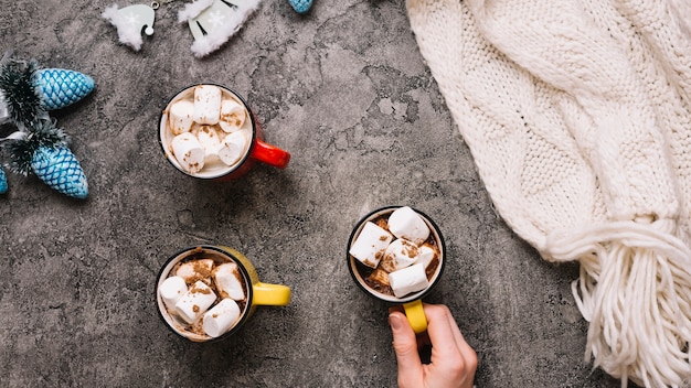 Mano che tiene la tazza con marshmallow vicino a tazze, giocattoli di Natale e sciarpa a maglia