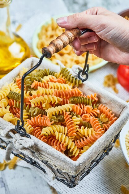 Mano che tiene un cesto di fusilli crudi con pasta assortita e verdura sul tavolo di marmo.