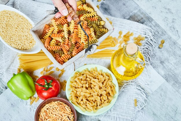 Mano che tiene un cesto di fusilli crudi con pasta assortita e verdura sul tavolo di marmo.