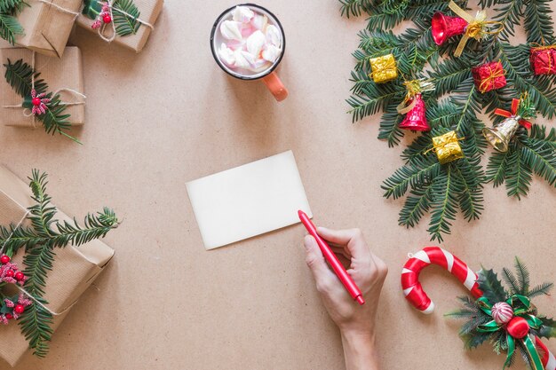 Mano con la penna vicino a scatole di carta vicino presenti, ramoscello e tazza di Natale