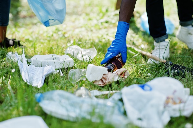 Mano dell'uomo afroamericano che raccoglie la bottiglia nei sacchi della spazzatura mentre pulisce l'area nel parco Africa volontariato di beneficenza e concetto di ecologia