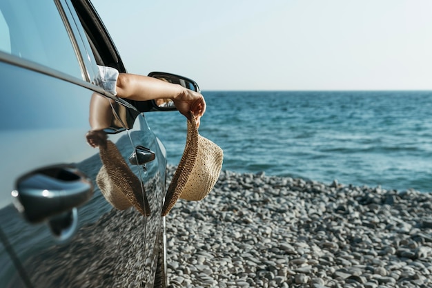 Mano della donna del colpo metà che pende dal finestrino della macchina e che tiene il cappello vicino al mare