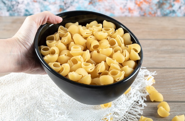 Mano femminile che tiene la ciotola piena di pasta secca sulla tavola di legno.