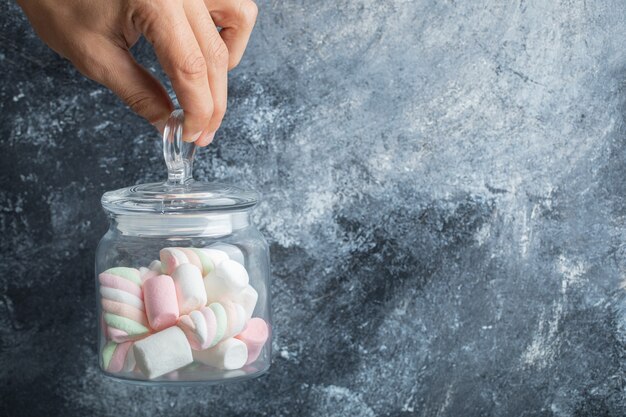 Mano femminile che tiene un barattolo di vetro di marshmallow colorati su sfondo di marmo