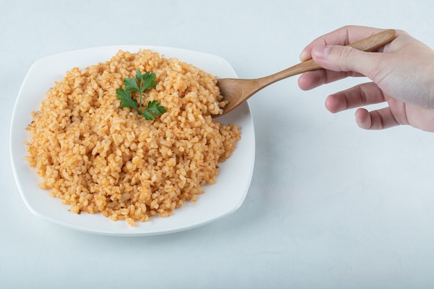 Mano femminile che tiene un cucchiaio di pilaf di bulgur sul piatto bianco.