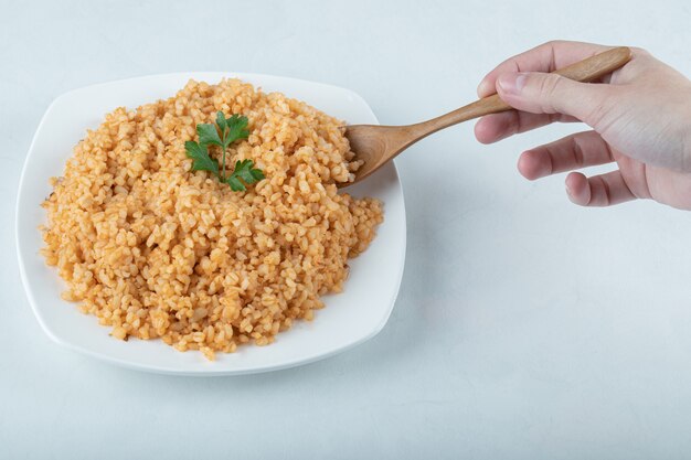 Mano femminile che tiene un cucchiaio di pilaf di bulgur sul piatto bianco.