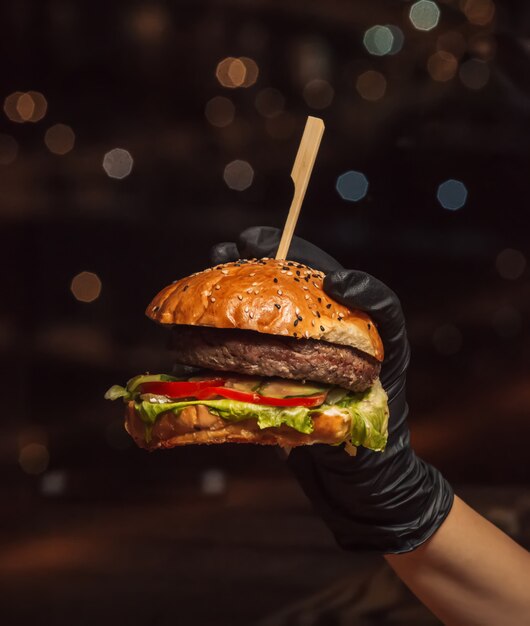 Mano nei guanti dell'hamburger che tengono l'hamburger di manzo nel fondo nero