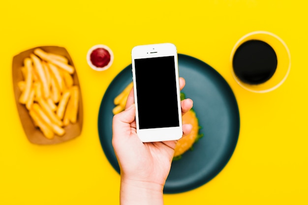 Mano piatta che tiene smartphone sul piatto con hamburger e patatine fritte