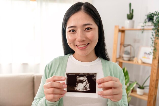 Mano sorridente asiatica della donna incinta che mostra le esplorazioni di ultrasuono a casa