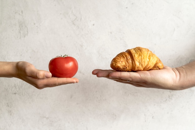 Mano umana che mostra croissant e pomodoro rosso davanti a sfondo concreto