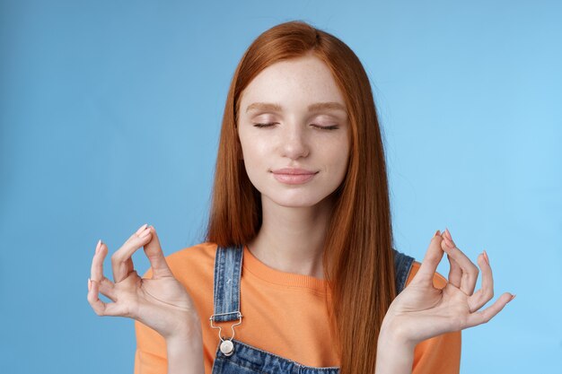 Mantieni la calma rossa ragazza rilassata rimanere sollevata positiva chiudere gli occhi sorridendo felice alzando le mani lateralmente loto mudra gesto pratica meditazione yoga fare esercizio di respirazione, sfondo blu.