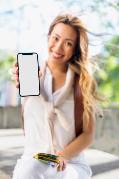 Mappa bionda sorridente della tenuta della giovane donna a disposizione che mostra schermo bianco in bianco mobile