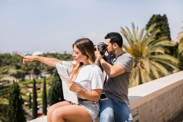 Mappa della tenuta della donna che indica dito che si siede con il suo ragazzo che prende foto sulla macchina fotografica nel parco