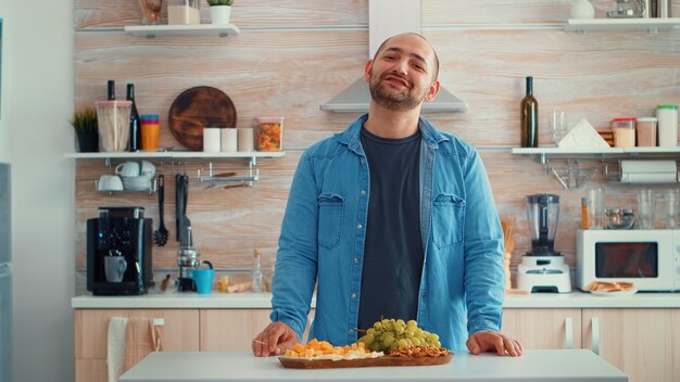 Marito chiacchiera in cucina mentre la sua famiglia prepara la cena in sottofondo. Ritratto felice sorridente giovane guardando la telecamera, colpo alla testa ritratto, mangiare formaggio, famiglia allargata intorno a lui