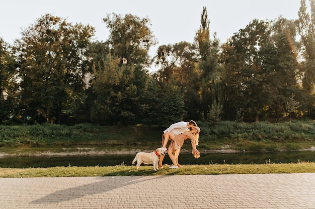 Marito e moglie che scherzano e ballano il tango nel parco vicino allo stagno mentre camminano con il Labrador.