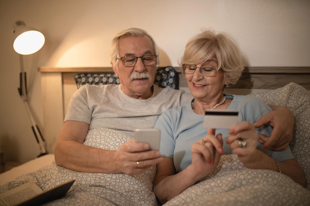 Marito e moglie maturi che usano smartphone e carta di credito mentre sono sdraiati a letto di notte