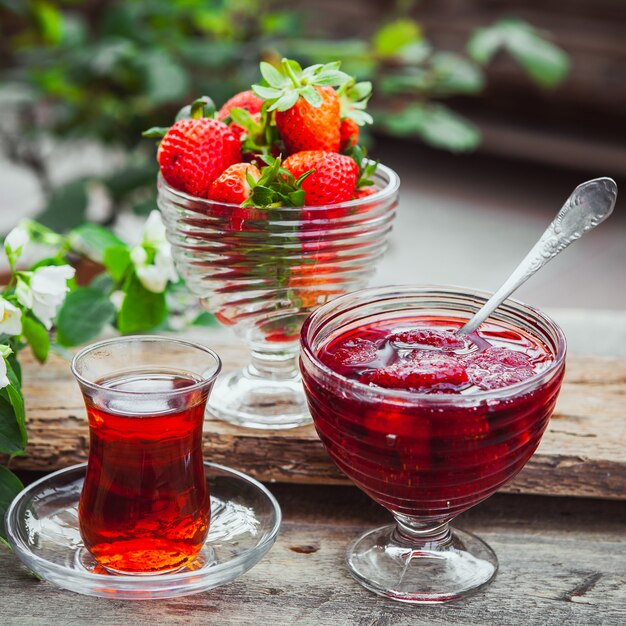 Marmellata di fragole in un piatto con cucchiaio, tè in vetro, fragole, primo piano della pianta sul tavolo in legno e cortile