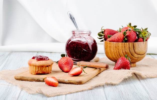 Marmellata e bigné di fragola di vista laterale con la fragola fresca in una ciotola e coltello di legno su fondo bianco