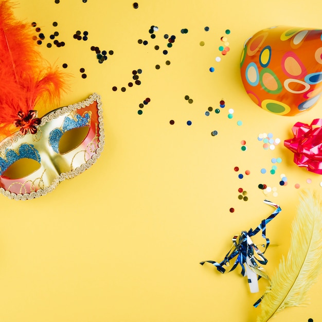 Mascherina della piuma di carnevale di travestimento con il materiale della decorazione del partito ed il cappello del partito sopra fondo giallo