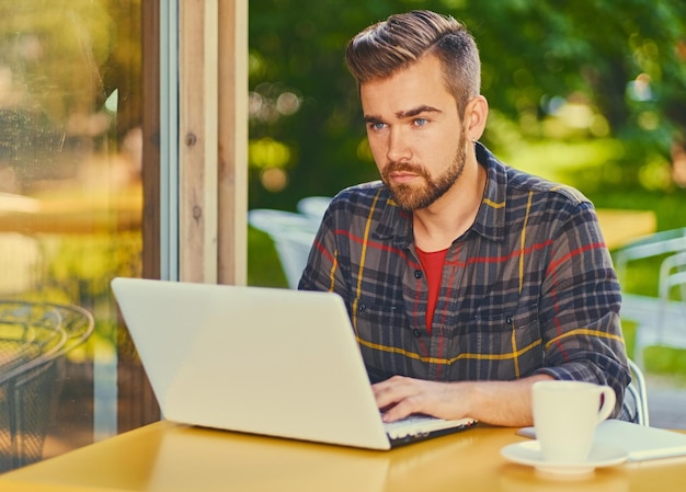 Maschio barbuto bello dei pantaloni a vita bassa che utilizza il computer portatile in un caffè.