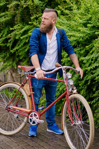 Maschio barbuto rosso vestito con una giacca blu e jeans su una bicicletta retrò in un parco.