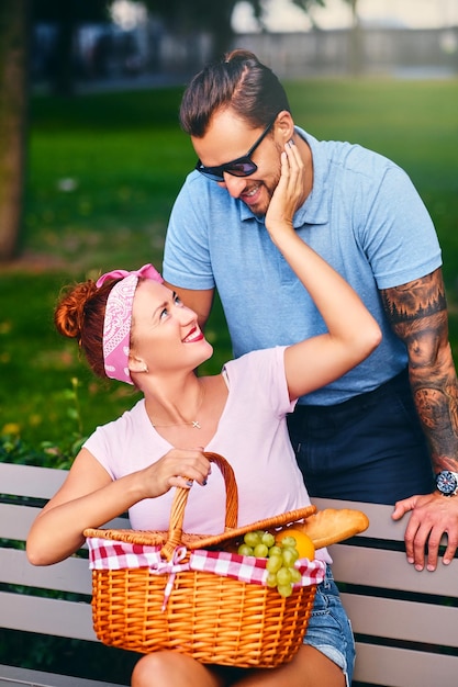 Maschio barbuto tatuato e femmina rossa stanno facendo un picnic su una panchina in un parco.