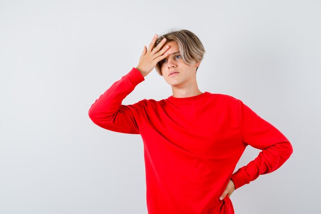 Maschio biondo teenager in maglione rosso con la mano sulla fronte, distogliendo lo sguardo e guardando pensieroso, vista frontale.