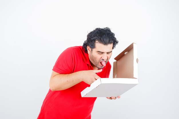 Maschio maturo in maglietta rossa guardando la scatola della pizza aperta mentre spuntava la lingua e sembra affamato, vista frontale.