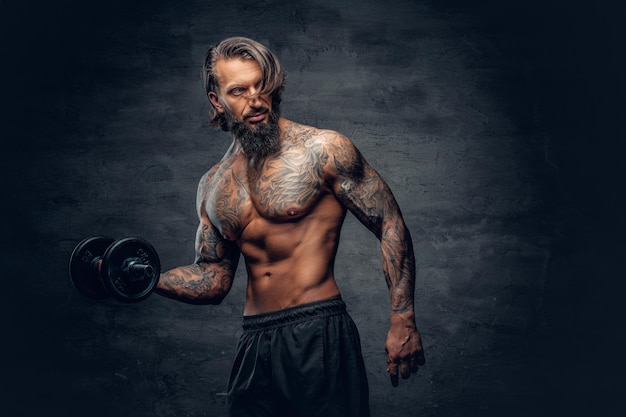 Maschio muscoloso e barbuto senza camicia con un tatuaggio sul busto che fa un allenamento bicipite con manubrio su sfondo grigio scuro.
