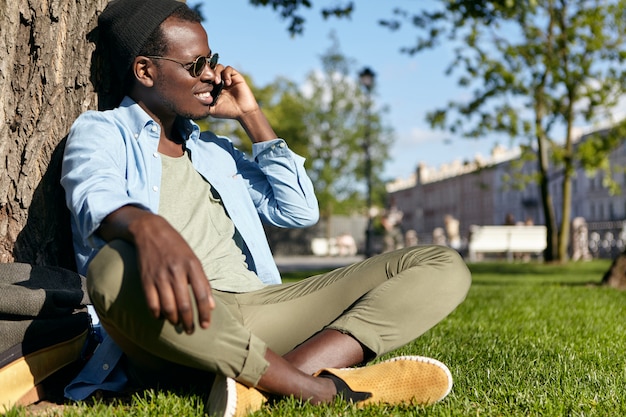 Maschio nero africano americano in abiti eleganti, seduto a gambe incrociate vicino all'albero al parco verde, chiacchierando sul suo cellulare, guardando da parte con espressione felice, ammirando splendido tempo all'aperto