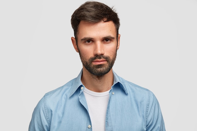 Maschio serio con la barba lunga dall'aspetto piacevole, ha i capelli scuri, setola, contempla qualcosa di importante, vestito con abiti alla moda, isolato su un muro bianco. Uomo europeo al coperto