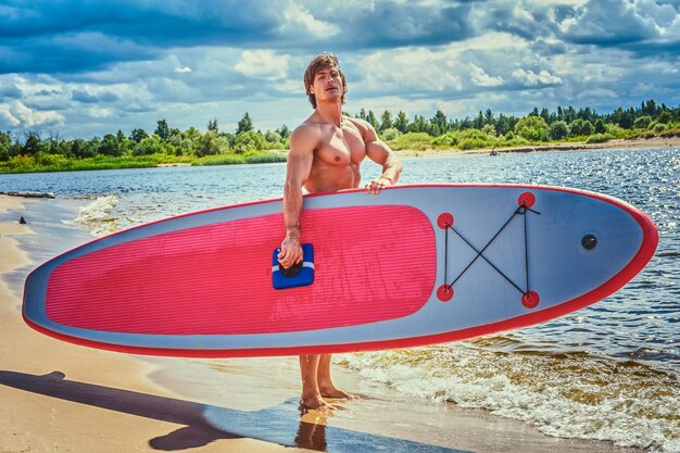 Maschio surfista senza camicia con un corpo muscoloso con la sua tavola da surf in spiaggia.
