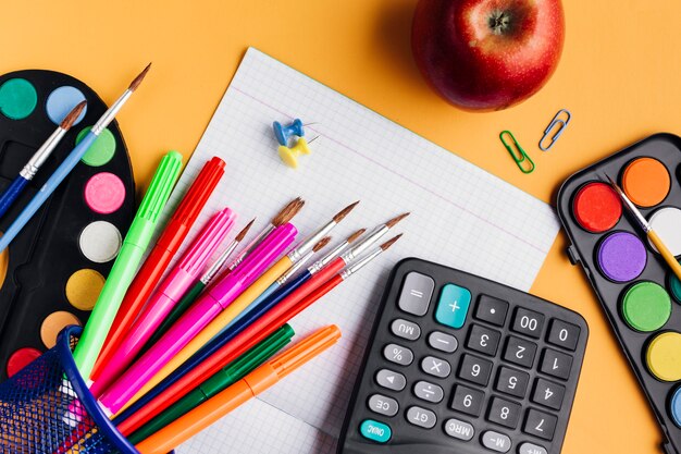 Materiale scolastico multicolore e mela rossa sparsi sulla scrivania gialla